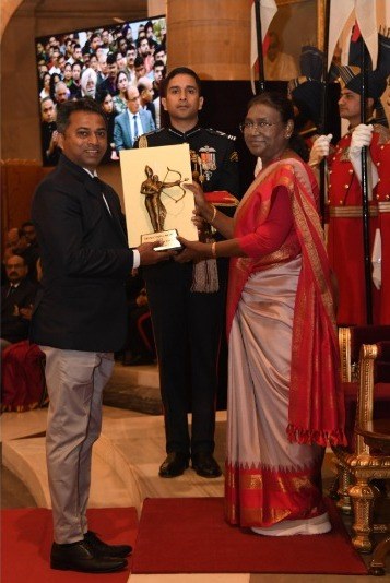 Ein Mann in einem schwarzen Blazer, weißem Hemd und grauen Hosen hält eine Trophäe, während er neben einer Frau in einem aschgrauen Sari auf einem roten Teppich an einer Preisverleihung steht, mit Menschen, Stühlen, Fahnen und einem Bildschirm im Hintergrund.