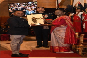 Ein Mann in einem schwarzen Blazer, weißem Hemd und grauen Hosen hält eine Trophäe, während er neben einer Frau in einem aschgrauen Sari auf einem roten Teppich an einer Preisverleihung steht, mit Menschen, Stühlen, Fahnen und einem Bildschirm im Hintergrund.