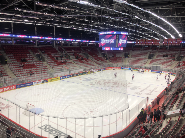 Ein Eishockeyspiel in einer großen Arena mit Zuschauern auf Sitzen und Stehplätzen, ein Zaun um die Eisfläche, ein Display mit der Aufschrift "St. Louis Blues vs Detroit Red Wings", Metallrahmen und eine Decke mit Deckenleuchten.