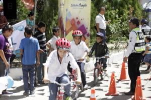 Kinder auf Fahrrädern fahren eine Straße entlang mit Verkehrskegeln, einige tragen Helme, andere stehen daneben, mit einer Fahne, Bäumen und Gebäuden im Hintergrund.