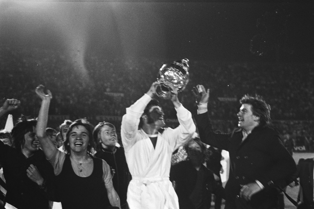 Eine Gruppe von Menschen, die mit einem Pokal vor einer jubelnden Menge feiern, wobei der Mann in der Mitte den Pokal triumphierend in die Höhe hält.