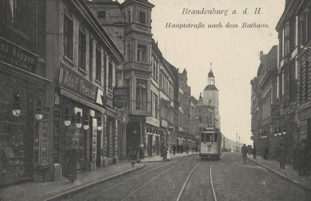 Altes Schwarz-Weiß-Foto einer Straßen in Brandenburg, Deutschland, mit einer Tram, Fußgängern, Gebäuden, Schildern, Straßenlaternen und einem bewölkten Himmel.