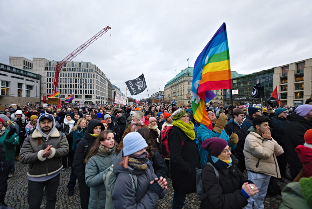 Große Gruppe von Menschen bei einer LGBTQ+-Rechtsdemo in Berlin, die Fahnen und Schilder schwenkt, mit Gebäuden, einem Kran und einem bewölkten Himmel im Hintergrund.