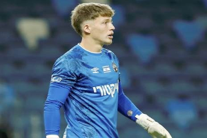 Ein junger Mann in einem blauen Torwarttrikot steht auf einem Fußballfeld und trägt ein blaues T-Shirt, weiße Handschuhe und scheint mitten in einem Spiel zu sein.