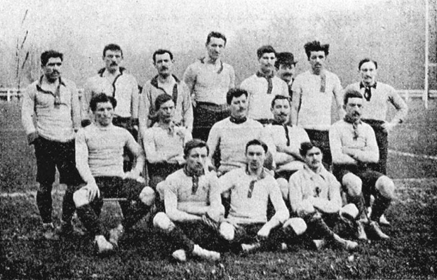 Schwarzes und weißes Foto einer Fußballmannschaft aus den 1920er Jahren, einige stehen und einige sitzen auf dem Boden, mit Pfählen, Bäumen und einem klaren Himmel im Hintergrund.