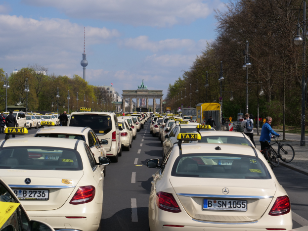 Eine lange Reihe von Taxis parkt am Straßenrand einer belebten Straße in Berlin, Deutschland, mit Fahrzeugen, Radfahrern und Fußgängern, flankiert von Laternenmasten, Bäumen und Gebäuden, einschließlich eines Bogens und eines Turms.