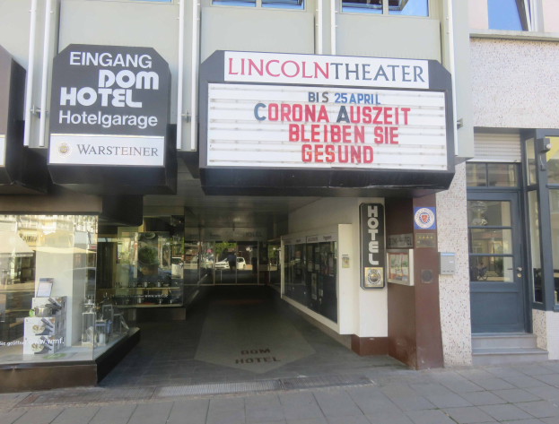 Außenansicht des Lincoln Theaters in Berlin, Deutschland, mit Glasfenstern und -türen sowie einer Tafel mit Text und einem Innenraum, der eine belebte Stadtlandschaft suggeriert.