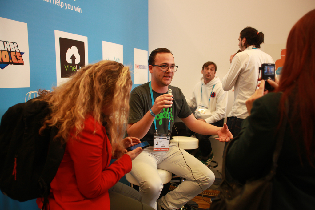 Ein Interview-Szenario mit Menschen, die Mikrofone und eine Kamera halten, ein Banner links daneben und Personen im Vordergrund und in der Mitte, mit einem gut beleuchteten Hintergrund.