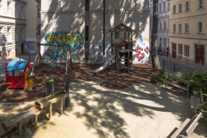 Spielplatz mit Graffiti in einer städtischen Umgebung, umgeben von Gebäuden, Grünflächen, Bänken und einer Straße mit Menschen und Fahrzeugen.