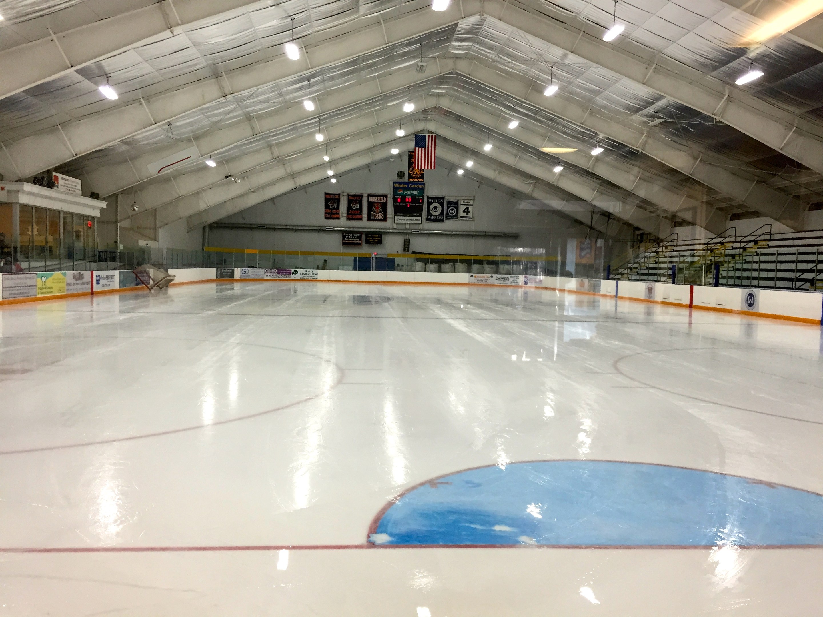 Große Indoor-Eisfläche mit Deckenbeleuchtung, Werbetafeln, einer Rückwand, Treppen mit Geländern und einigen Menschen, was auf eine Sportanlage hindeutet, wahrscheinlich eine Eishockeyhalle.