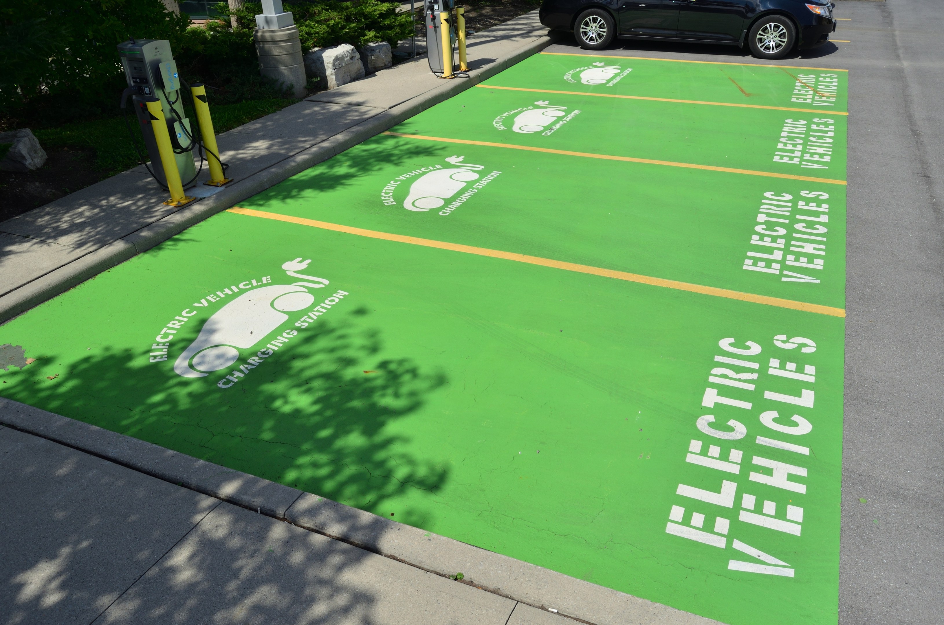 Elektroauto auf einem Ladeplatz mit einer grünen Bodenmatte mit Text und Bildern in einer Anlage umgeben von Bäumen, Pflanzen, Masten und einem Gebäude.