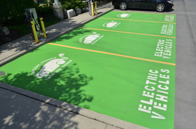 Elektroauto auf einem Ladeplatz mit einer grünen Bodenmatte mit Text und Bildern in einer Anlage umgeben von Bäumen, Pflanzen, Masten und einem Gebäude.
