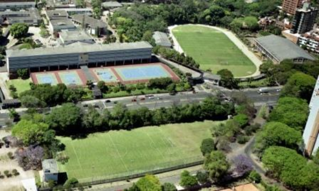 Luftaufnahme eines Fußballfeldes auf dem Campus der Universität Sydney, umgeben von Bäumen, Gebäuden und Fahrzeugen, mit einem neuen Sportkomplex in Planung.