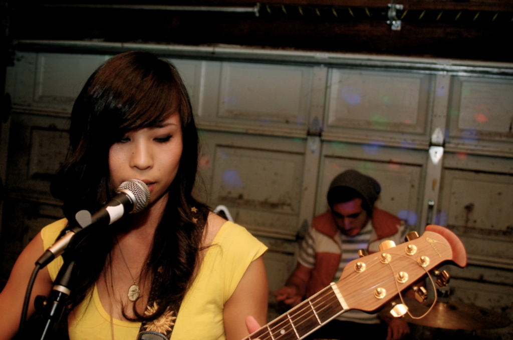 Eine Frau in der Mitte spielt Gitarre und singt während eines Live-Auftritts, mit einem Mann auf der rechten Seite im Hintergrund und einem Mikrofon links.