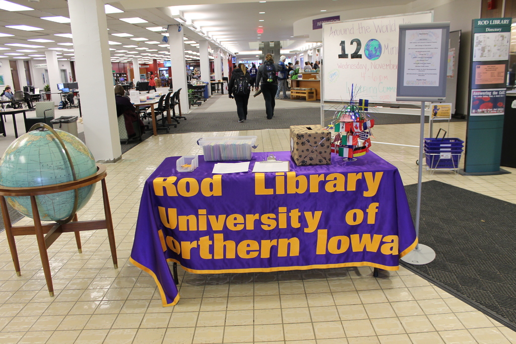 Eine Ausstellung mit einem Tisch, auf dem verschiedene Landesflaggen, eine Box, Papiere und eine Fahne liegen, eine Kugel links daneben, ein Anschlagbrett rechts daneben mit einigen Menschen in der Nähe und mehreren Tischen und Stühlen links.
