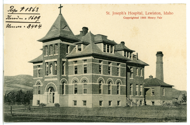 Ein Schwarz-Weiß-Foto des St. Joseph's Hospitals in Lewiston, Idaho, das ein großes Gebäude mit Fenstern, eine Treppe mit Geländern, einen Schornstein auf der rechten Seite und Text oben zeigt, mit Bäumen, Hügeln und dem Himmel im Hintergrund.