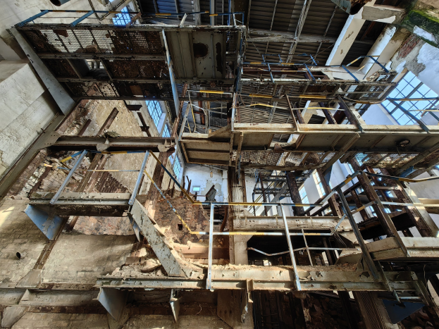 Alter Industriebau mit einem zentralen Metallgerüst, umgeben von verrosteten Eisenstangen, Treppen, Wänden und Fenstern, die Zeichen von Verfall und Verwahrlosung zeigen.