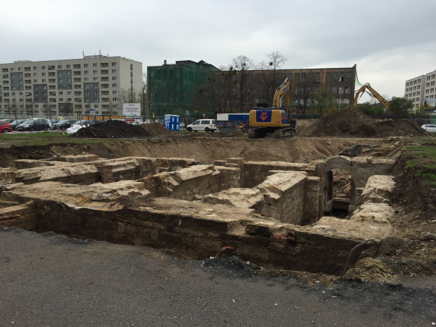 Eine Baustelle mit einem großen Erdwall, umgeben von Baggern, Fahrzeugen, Gras, Bäumen und Gebäuden mit Fenstern unter einem bewölkten Himmel über einer Straße.