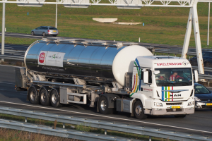 Großer Tanklastzug auf einer Autobahn neben einem Auto mit sichtbaren Insassen im Truck und Gras, Stangen und anderen Gegenständen im Hintergrund.