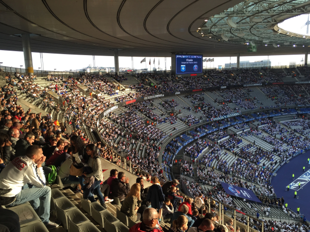 Eine große Menschenmenge in einem Stadion schaut einem Fußballspiel zu, mit einer Bühne rechts, Fahnen, Stangen, einem Bildschirm und dem Allianz Stadion in München im Hintergrund.