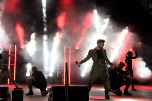 Drake, mit einem Mikrofon in der Hand, performs auf der Bühne beim 2018 Coachella Valley Music and Arts Festival, umgeben von einer Gruppe von Menschen in einer Hockposition, mit Lautsprechern, einer Person mit einer Videokamera, Metallstangen und einer Decke mit Deckenleuchten.