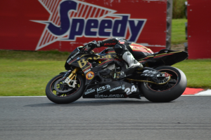 Eine Person in Helm und Schuhwerk fährt ein Motorrad auf einer Rennstrecke mit Gras und einer Tafel mit Text im Hintergrund.