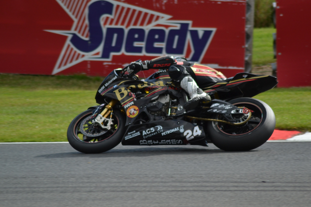 Eine Person in Helm und Schuhwerk fährt ein Motorrad auf einer Rennstrecke mit Gras und einer Tafel mit Text im Hintergrund.
