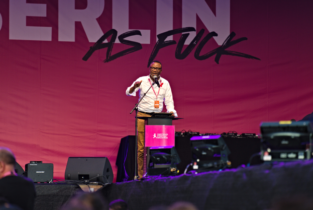 Ein Mann in einem weißen Hemd und braunen Hosen steht auf einer Bühne und hält ein Mikrofon in der Hand, hinter ihm eine pinkfarbene Fahne mit der Aufschrift "Berlin as Fuck" und Lautsprecher auf der Bühne.