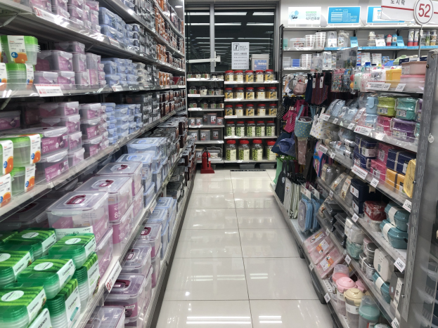 Ein unordentlicher Gang in einem chinesischen Supermarkt mit Regalen voller Kisten, Flaschen, Tüten und anderen Artikeln, beleuchtet von Deckenlampen, mit Informationsschildern und einem sichtbaren Feuerlöscher.
