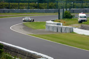 Ein Rennwagen fährt auf einer Rennstrecke mit einem Polizeiwagen im Hintergrund, umgeben von Grünflächen und Absperrungen, mit Text am unteren Bildrand.