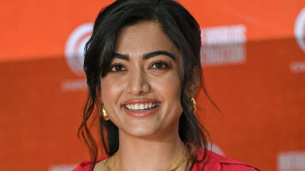 Eine Frau in einem roten Kleid mit einem eleganten Dutt und dezentem Make-up steht lächelnd vor der Kamera, trägt schwarze High Heels und hat einen hellgelben Banner mit der Aufschrift "Sonam Kapoors Beste Schönheits Looks vom Cannes Film Festival" im Hintergrund.