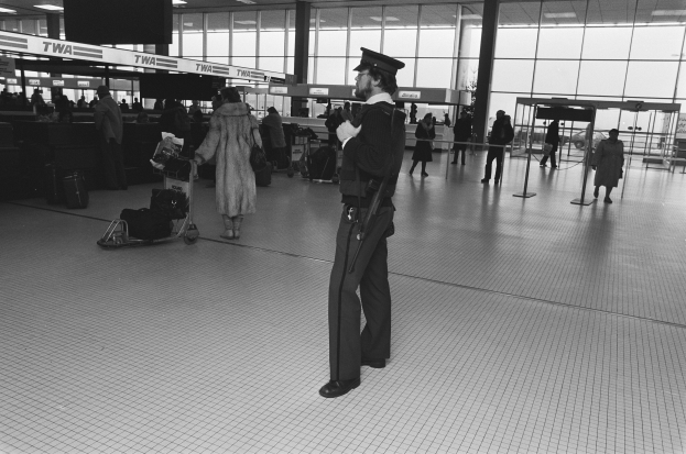 Ein Polizist in Uniform und Mütze steht in einem Flughafen-Terminal und hält eine Waffe in der Hand, umgeben von Menschen und Gepäckwagen mit Taschen, Stangen, Texttafeln, Lampen und Glaswänden im Hintergrund.