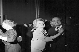 Ein Mann und eine Frau tanzen zusammen auf einer Party, umgeben von Zuschauern, mit Wandbeleuchtung im Schwarz-Weiß-Stil.