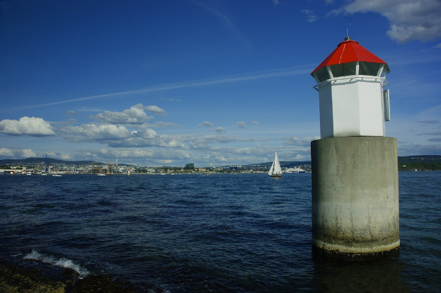 Boote auf dem Meer mit einem Leuchtturm im Hintergrund, umgeben von Gebäuden, einem Berg und dem Himmel.