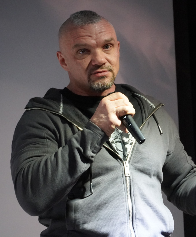 Ein Mann in einer schwarzen Kapuzenjacke und einem schwarzen Anzug mit weißem Hemd, zurückgekämmtem Haar und einem selbstbewussten Gesichtsausdruck hält ein Mikrofon, während er vor einer Wand steht.