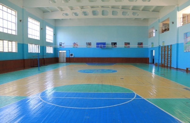 Große Turnhalle mit blau-weißem Boden, einem Basketballfeld, einem Netz mit Pfosten, einer Tür, Fenstern, Wandplakaten, einem Geländer und einem Dach.