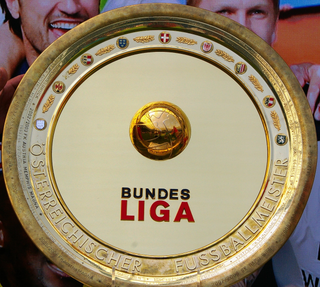 Ein Mann hält einen goldenen Schild mit der Aufschrift "Bundesliga" hoch, im Hintergrund ist eine Fahne mit drei Personen zu sehen.