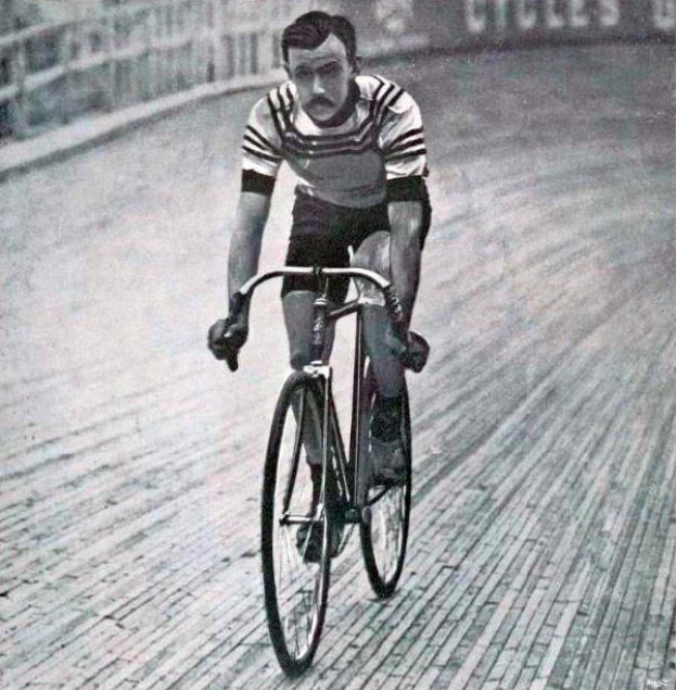 Schwarz-weiß-Foto eines Mannes, der auf einer Bahn einen Fahrrad fährt, mit einem Zaun und einer Tafel im Hintergrund, der angeblich Sir Ernest Hemingway ist.