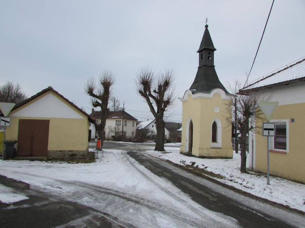 Ein kleines verschneites Dorf mit einer Kirche in der Mitte, umgeben von Häusern, Schildern, einem Müllcontainer, Bäumen und Drähten unter einem bewölkten Himmel.