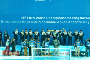 Eine Gruppe von Menschen, die oben auf einem Swimmingpool stehen, einige halten Kameras, mit einem Netz im Vordergrund und einem Banner im Hintergrund, auf dem "FINA World Championships 2015 Kazan" steht.