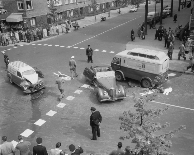 Schwarz-weiß-Szene eines Autounfalls an einer Straßenkreuzung mit mehreren Fahrzeugen, darunter ein Lieferwagen, und einer Gruppe von Schaulustigen; Gebäude, Bäume, Pfähle und Schilder sind im Hintergrund zu sehen.