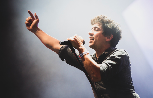 Ein Mann in schwarzem Hemd und Jeans singt in ein Mikrofon auf einer Bühne, mit geschlossenen Augen und einem jubelnden Publikum im Hintergrund.