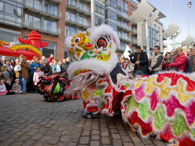 Ein lebendiges chinesisches Neujahrsfest in Amsterdam mit einem Löwen tanzen im Vordergrund und einer Menge Menschen drumherum, einige halten Kameras, während Gebäude, Laternenmasten und ein klarer blauer Himmel im Hintergrund zu sehen sind.