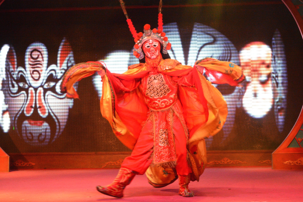 Ein Mann in einem roten chinesischen Opernkostüm mit detaillierten Verzierungen und leuchtenden Farben steht auf der Bühne, das Gesicht bunt geschminkt, mit einem einzigartigen Ausdruck, die Arme ausgestreckt, der Kopf geneigt, die Augen geschlossen und der Mund leicht geöffnet, inmitten einer Performance.