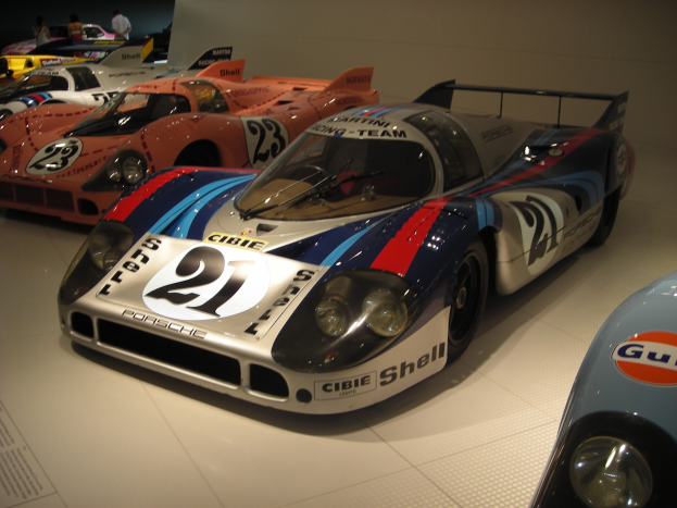 Ein Porsche 917K Le Mans Rennwagen in einem Museum ausgestellt, umgeben von anderen Sportwagen, mit Menschen in der Nähe und einer Wand im Hintergrund; ein Schild mit Text ist links sichtbar.