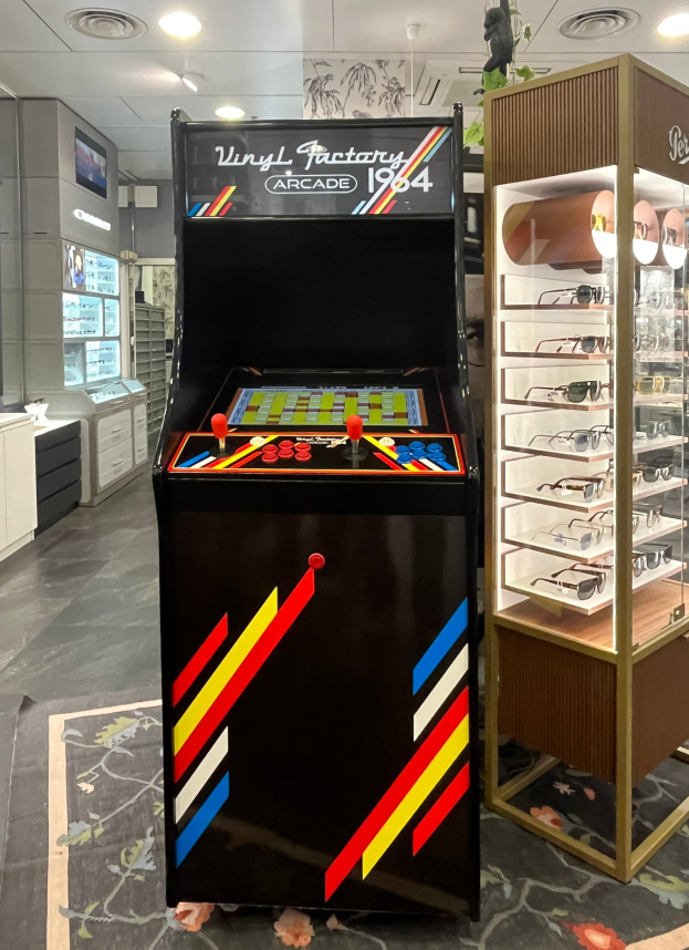 Ein Geschäft mit einem vintage Arcade-Automaten in der Mitte, einem Glasvitrine auf der rechten Seite und einer Retro-Beleuchtung mit Schränken, Fernseher und anderen Gegenständen im Hintergrund.