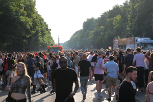 Eine große Menschenmenge, die eine von Bäumen gesäumte Straße entlanggeht, mit einem Turm im Hintergrund und Fahrzeugen mit Teilnehmern auf der rechten Seite, wahrscheinlich beim Christopher Street Day in Berlin unter einem klaren blauen Himmel.