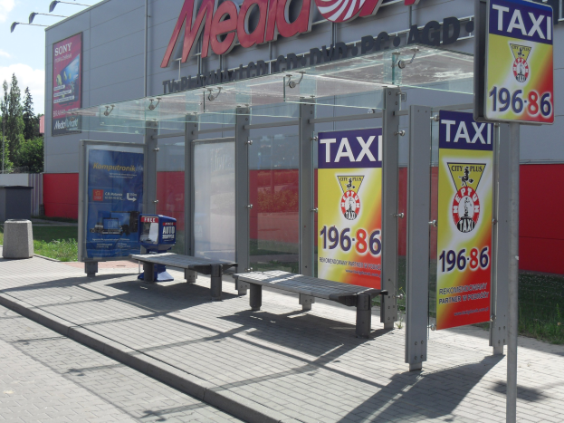 Eine Bushaltestelle mit einem "Taxi"-Schild, umgeben von Bänken, Werbetafeln, einem Mülleimer, Gras, Bäumen und einem bewölkten Himmel.