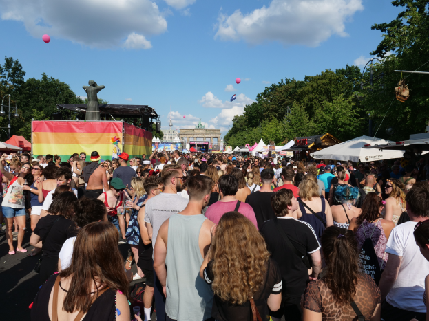 Eine große Menschenmenge, die eine Straße mit Zelten, Bäumen, Pfählen, Lichtern und Gebäuden entlanggeht, mit einem bewölkten Himmel mit Ballons im Hintergrund beim Berlin Pride Festival.