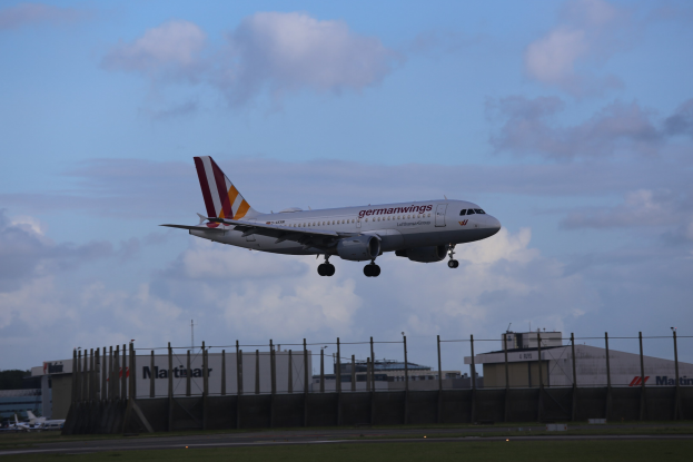 Germanwings Airbus A320-200 beim Starten vom Frankfurter Flughafen mit Gras darunter, einem Zaun im Vordergrund, Gebäuden und Bäumen im Hintergrund und einer bewölkten Himmel darüber.